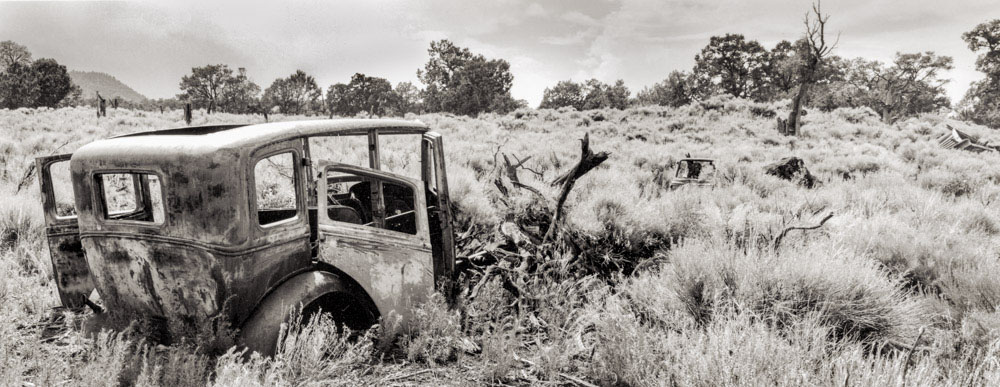 Abandoned vintage car rusting in a dry, overgrown field with trees in the background.