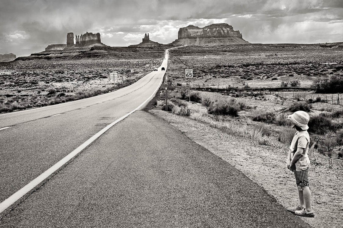 Young girl stands by side of highway with desert landscape.