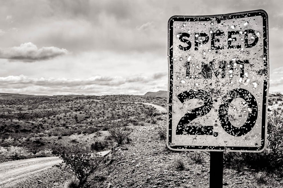 A weathered and bullet-riddled speed limit sign reading 20 stands beside a dirt road winding through a barren, rocky desert landscape under a cloudy sky.