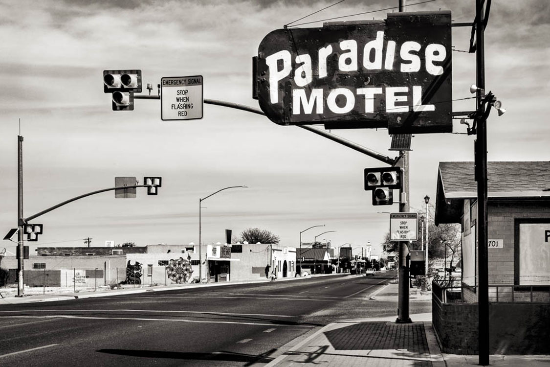 A black and white photo of a vintage “Paradise Motel” sign over a quiet, empty street with buildings and traffic lights in the background.