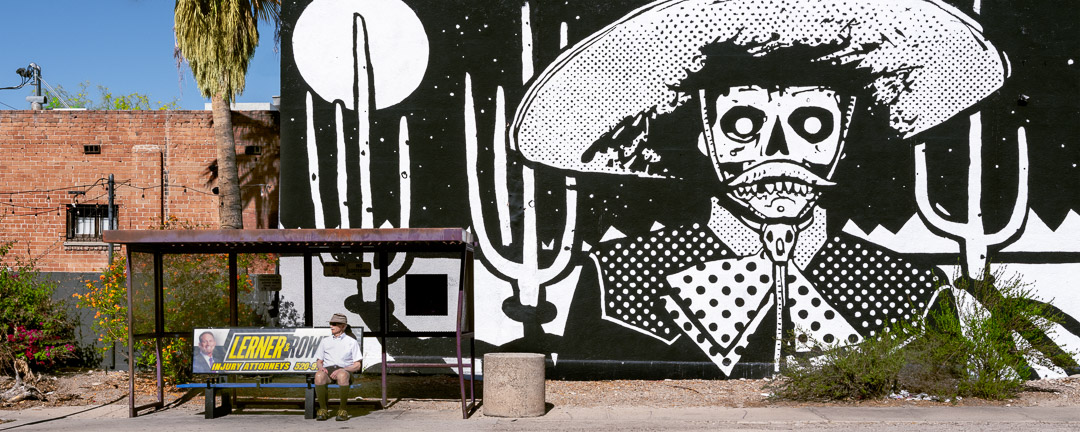 Urbanwalls 7. 250427 AZ _33_28 Panoramic image of B&W mural of skeleton cowboy in front of saguaro cactus and moon. Mural is behind man sitting at bus stop. Tuscon.